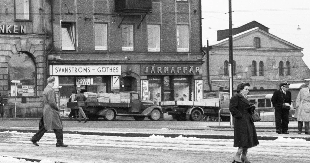 Gatuscen från Göteborg under 1900-talet med människor på snöig väg, butiker med skyltar för Svanströms och Järnaffär, klassicistisk byggnad i bakgrunden.